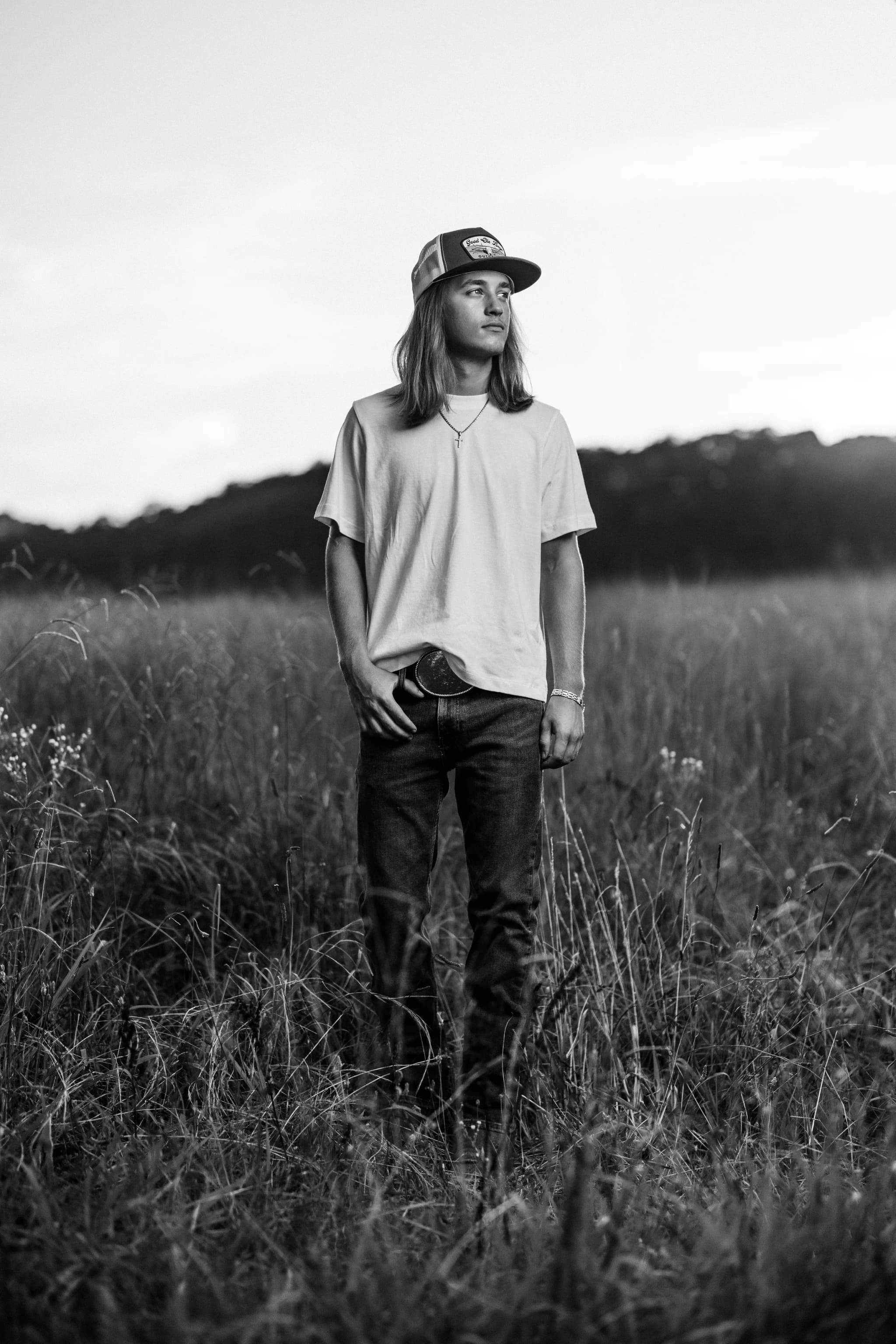 Black-and-white senior portrait of a long-haired guy in a trucker cap standing in a tall grass field