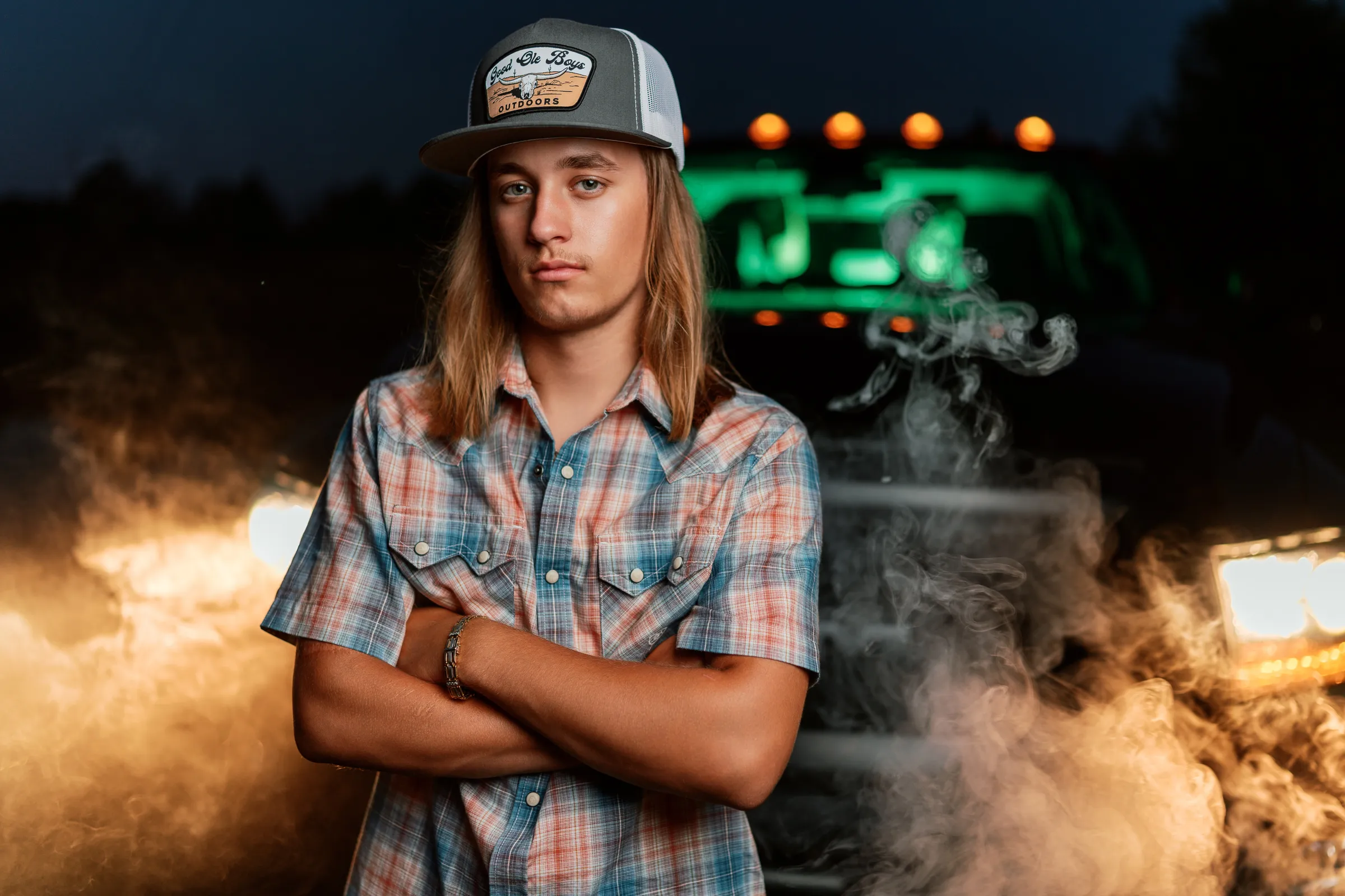 Senior in a plaid western shirt, arms crossed in front of his truck's glowing lights and drifting smoke at night
