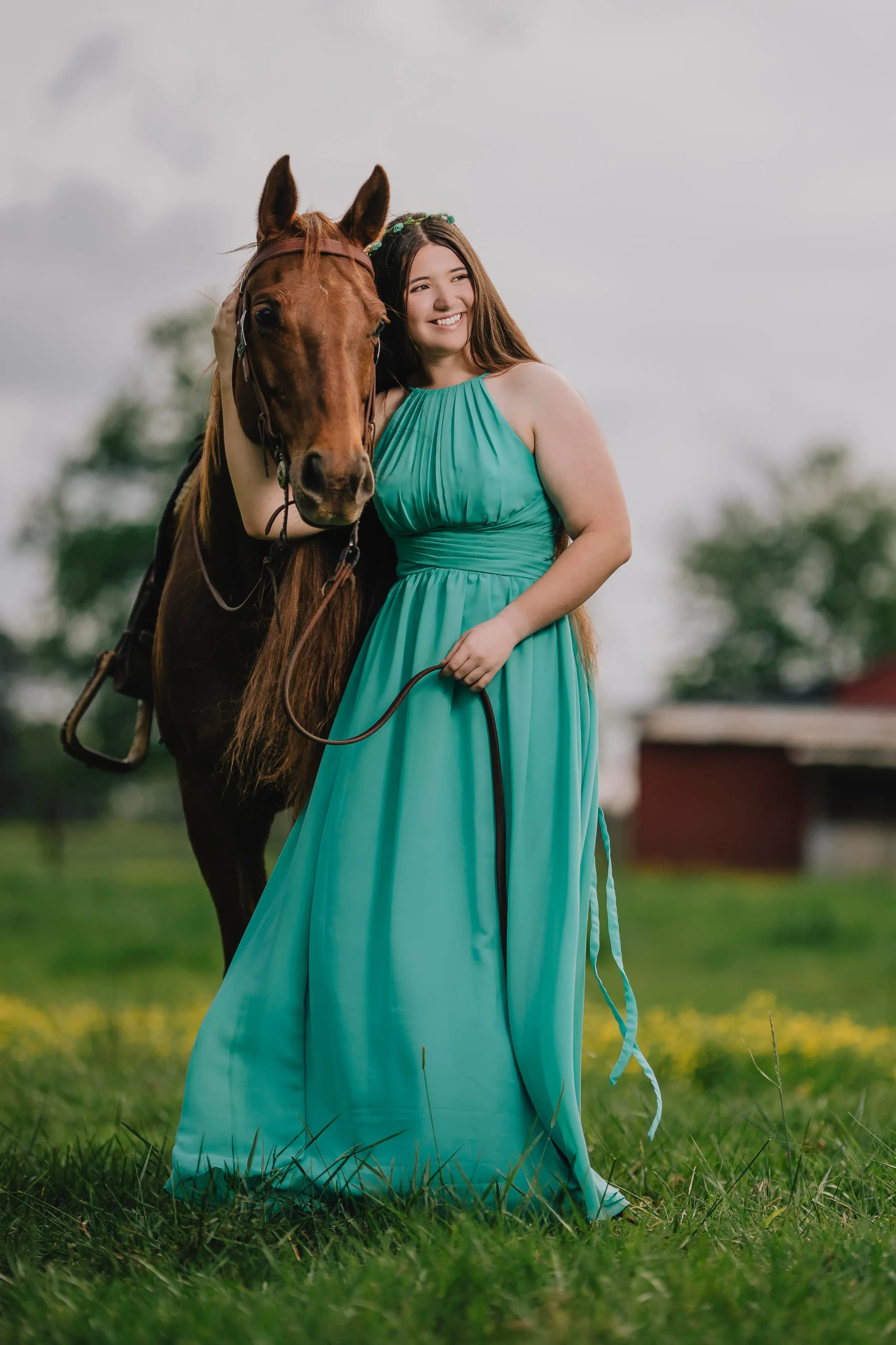 Girl in a flowing teal gown standing with her chestnut horse in a farm pasture