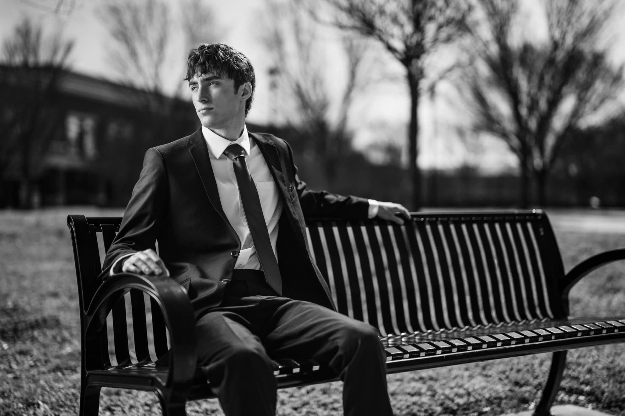 Senior in a black suit seated on an iron park bench, gazing off in dramatic black and white