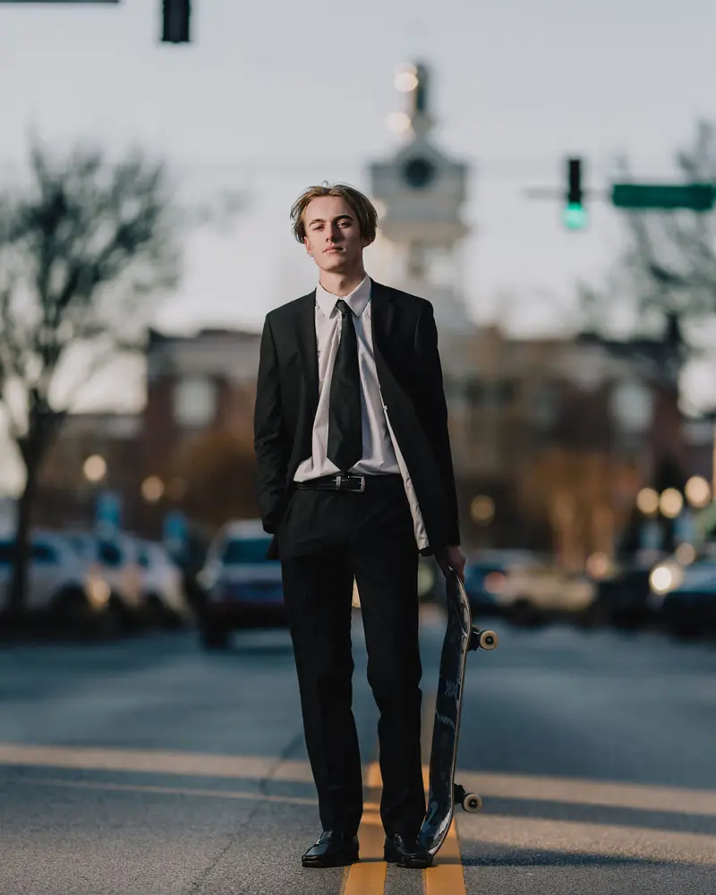Senior guy in a black suit holding a skateboard in the middle of a historic downtown street at dusk