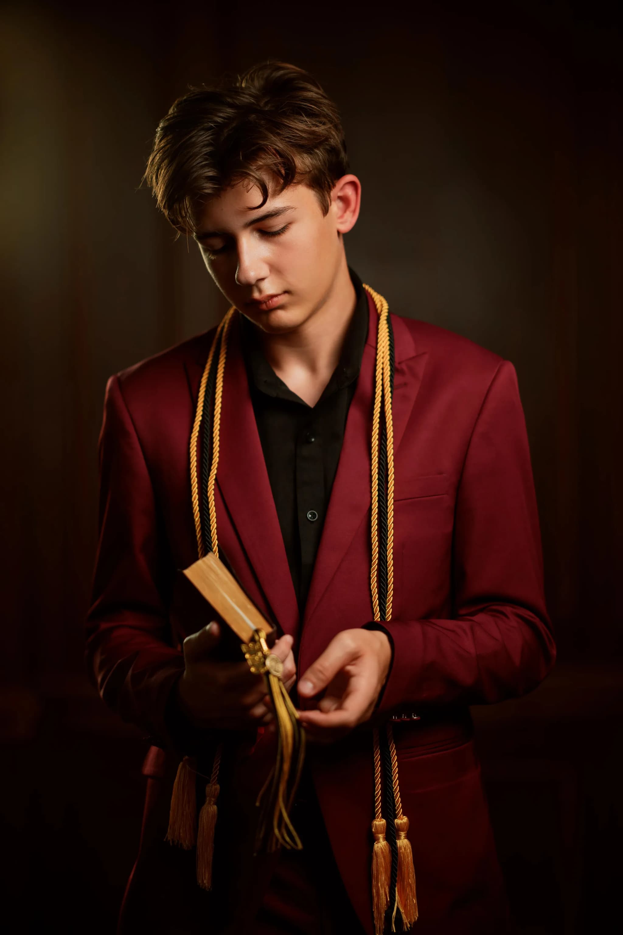 Senior in a burgundy suit with gold honor cords, lit like an old master painting