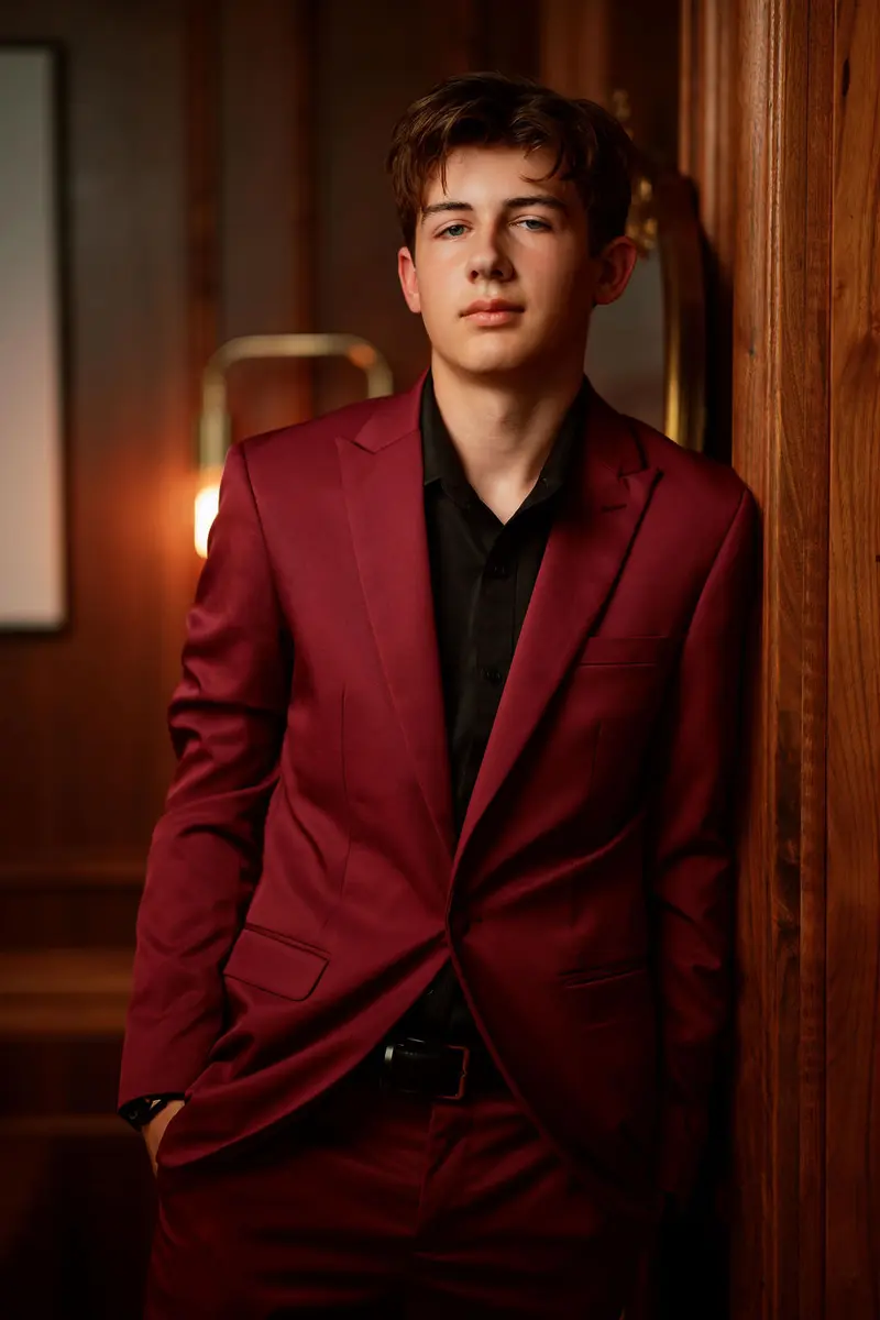 Senior in a burgundy suit leaning in a wood-paneled doorway under warm lamplight