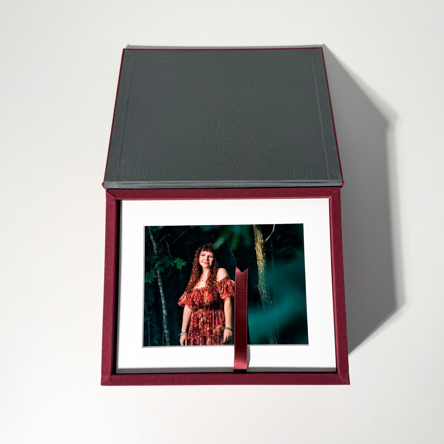 Burgundy linen keepsake box open, revealing a matted senior portrait tied with a ribbon
